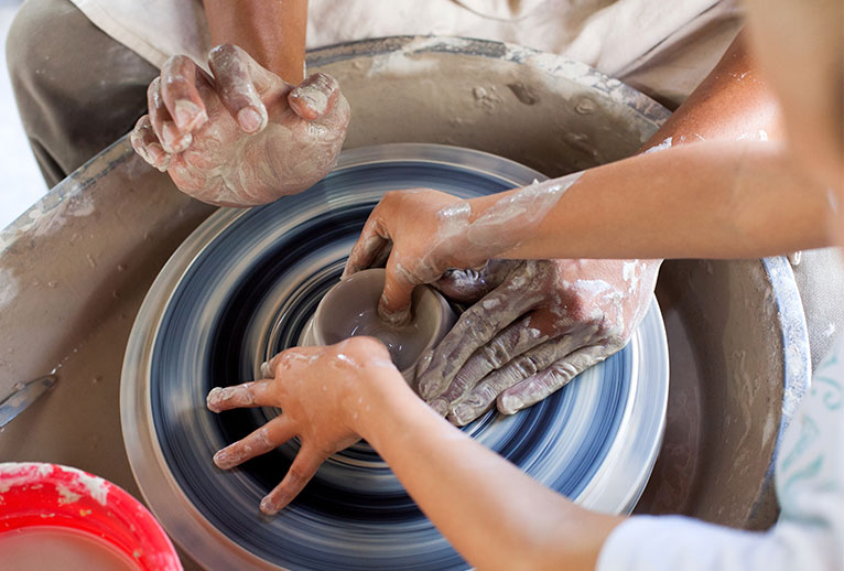 A potter on a potter's wheel receiving instruction on how to mold clay with their hands.