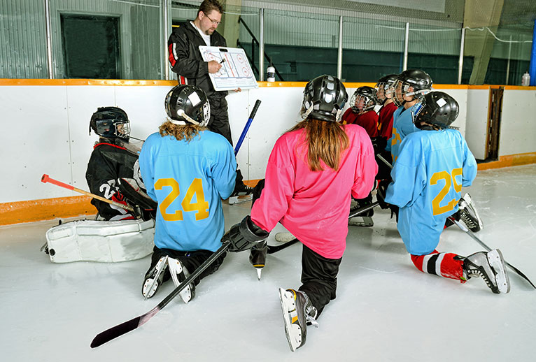 Group of youth geared up for ice hockey kneeling on the ice while their coach shows the schematics of a play.