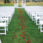 A trail of red rose pedals lay in an aisle of green grass leading to a white gazebo with white chairs on either side.