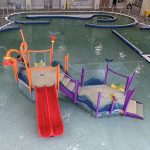Play feature in the Activity Pool inside Ridge Recreation Center showing colorful apparatuses and slides with the full pool and the lazy river in the background.