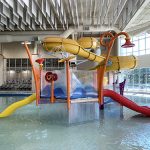 Play feature in the Activity Pool inside Ridge Recreation Center showing colorful apparatuses and slides with the large, yellow flume slide in the background.