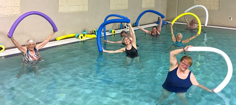 Participants standing in a pool holding pool noodles over their heads while stretching to their side.