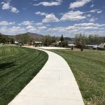 Sidewalk leading into Trappers Glen Park showing green grass, park shelter, basketball pad, playground and mountain views on a blue sky day.