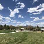 Wide view of Trappers Glen Park showing green grass, sidewalks, park shelter, basketball pad, playground and mountain views on a blue sky day.