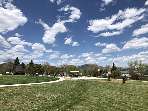 Wide view of Trappers Glen Park showing green grass, sidewalks, park shelter, basketball pad, playground and mountain views on a blue sky day.