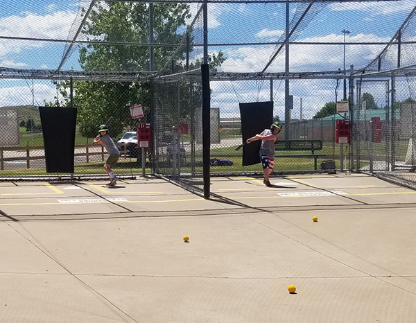 Two boys swinging bats at balls inside a batting cage.