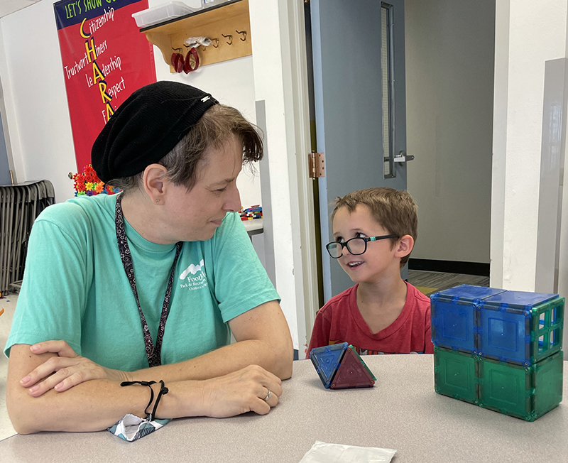 A young boy playing with blocks with a smile on his face looking up at his camp counselor.
