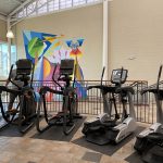 Cardio equipment lined up in the cardio weight room with a colorful mural on the wall in the background.