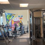 Cardio equipment area with lifting machines and exercise bikes with a colorful mural on the wall in the background.
