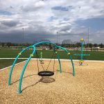 Pieces of playground equipment with sports fields nearby in the background.