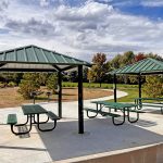 Two small park shelters with picnic tables underneath with new entry way, sidewalk and grills in the background.