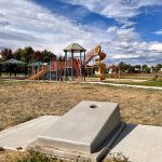 Play features in a park showing a cornhole game affixed on a cement pad and a playground in the background.