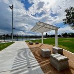 Sidewalk view with seating and gazebo shade structure leading toward fenced in pickleball courts.