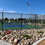 Sidewalk running alongside fenced in pickleball courts while people play the sport.