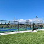 Sidewalk running alongside fenced in pickleball courts showing benches outside the fences to watch players.