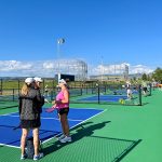 A group of women standing on a pickleball court chatting while games of pickleball occur on courts behind them.