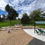 New stone seating wall bordering a portion of the park and playground with a park shelter and picnic bench on the side.