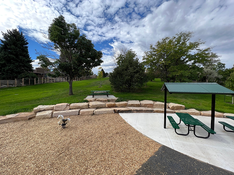 New stone seating wall bordering a portion of the park and playground with a park shelter and picnic bench on the side.
