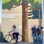 Three people pose on an interactive art piece painted on a wall showcasing a tree with a swing and a bike