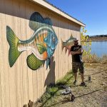 Artist Patrick Maxcy posting with the blue gill fish he painted on the side of a shed