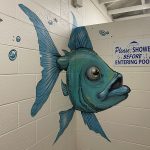 A blue fish painted on a wall near a sign that reads please shower before entering pool