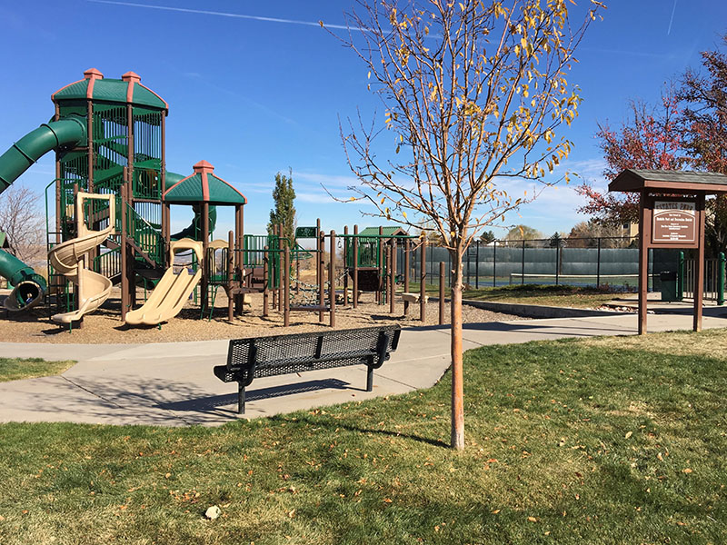 Park view of a tree and park bench overlooking a playground, park entry sign, picnic shelter and tennis court