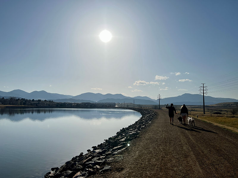 A trail made of crusher fines next the lake with mountain views showing two people walking dogs on leash