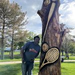 Artist Bongo Love poses with his tree carving artwork showcasing several different sports themed equipment.