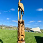 Tree carving art of a blue heron and fish with a backdrop of a lake, blue skies, amphitheater and mountain views.