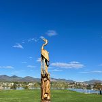 Tree carving art of a blue heron and fish with a backdrop of a lake, blue skies and mountain views.