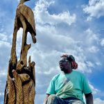 Artist Bongo Love sitting next to his tree carving artwork of a blue heron.