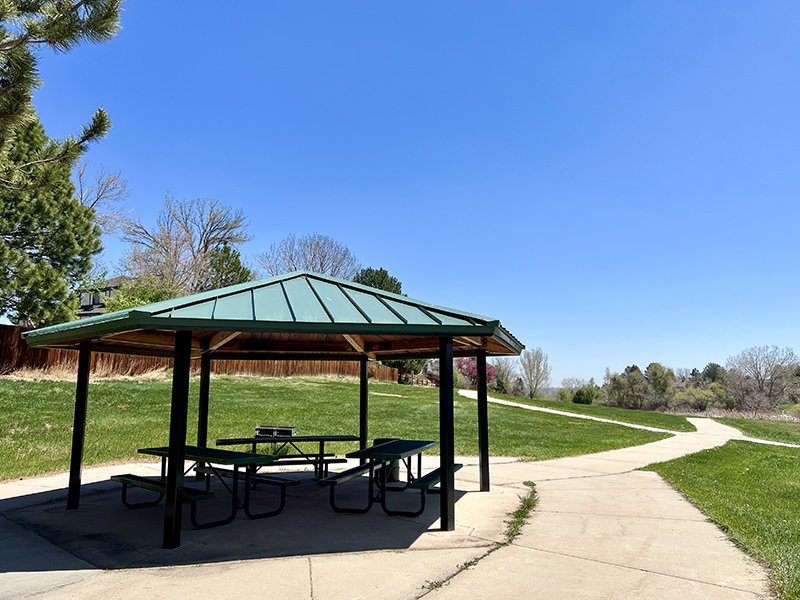 A quaint park shelter with picnic benched placed underneath alongside concrete trails and green grass