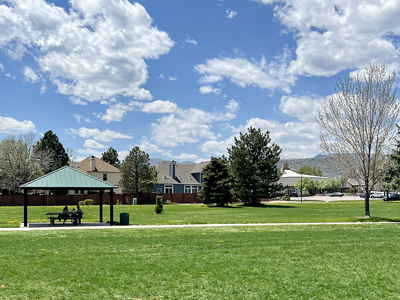 Green grass, a concrete trail, trees and a shelter with a picnic bench underneath