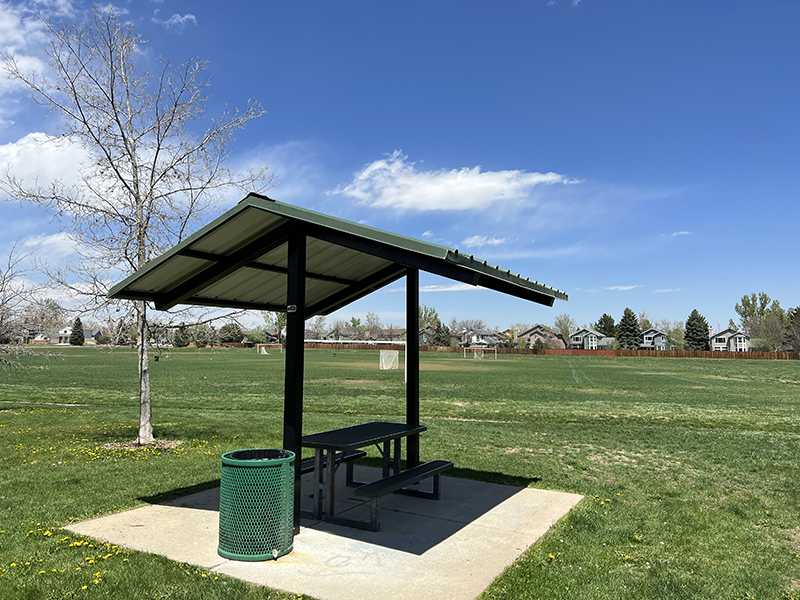 A small park shelter with a picnic bench underneath, trash can, trees and green grass with soccer fields