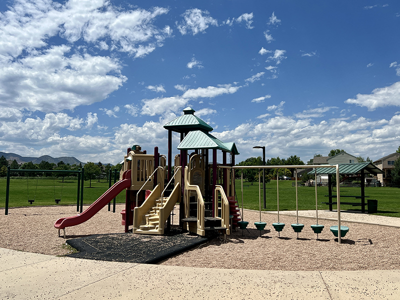 A playground with two slides, climbing apparatuses, swings and a park shelter and table with green grass and mountains in the background