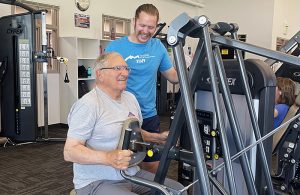 A fitness instructor guides a man using a piece of a fitness equipment.