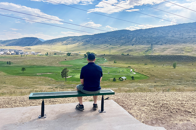 A man sitting on a bench off the Dutch Creek Trail overlooking the Meadows Golf Club and foothills in the background.