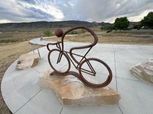 A piece of public art at the Dutch Creel Trail head located next to Ridge Recreation Center. The piece is titles "Pathfinder" and is a metal sculpture of a cyclist. 