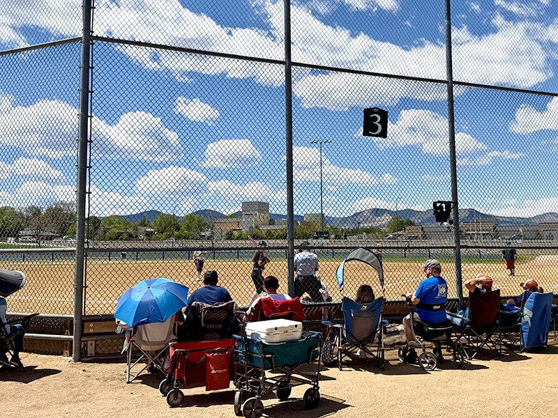 Onlookers watching from behind a fence while a softball game is in play.